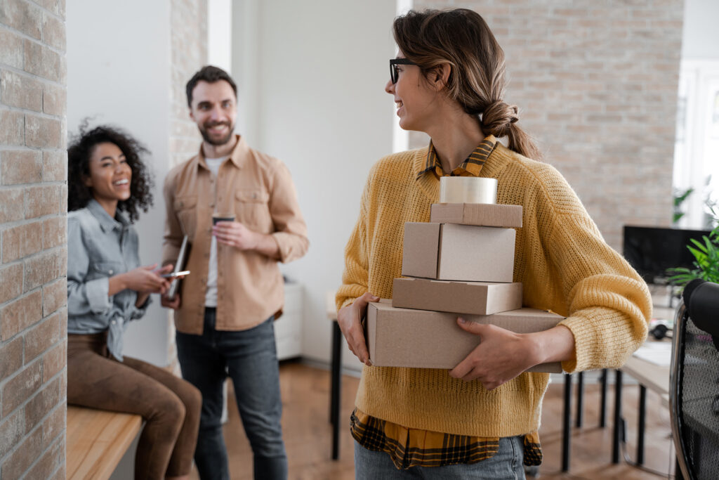 Happy woman holding boxes with smiling friends in the background, celebrating teamwork and success.